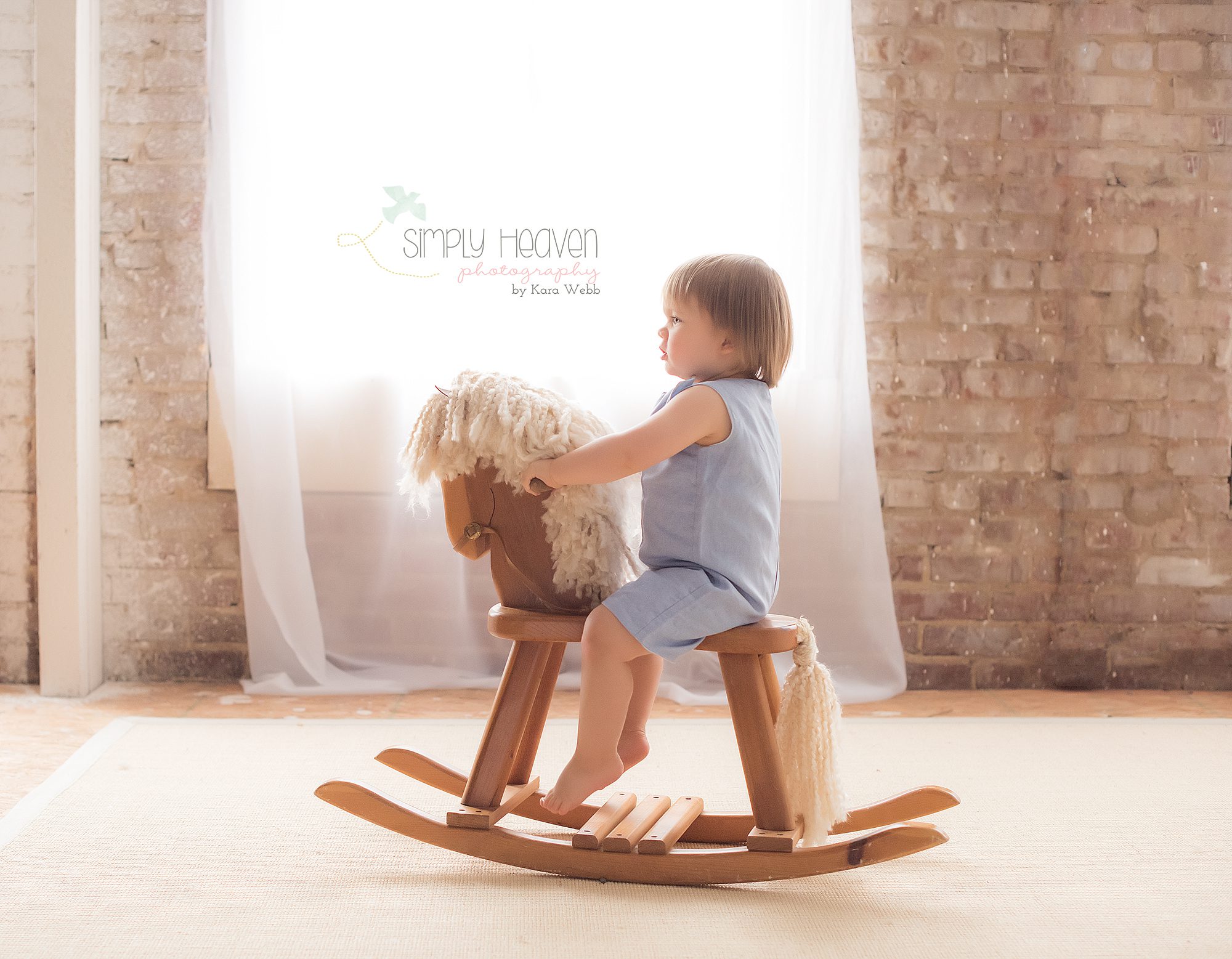 18 month baby boy in a blue romper rocking on a horse in front of a window