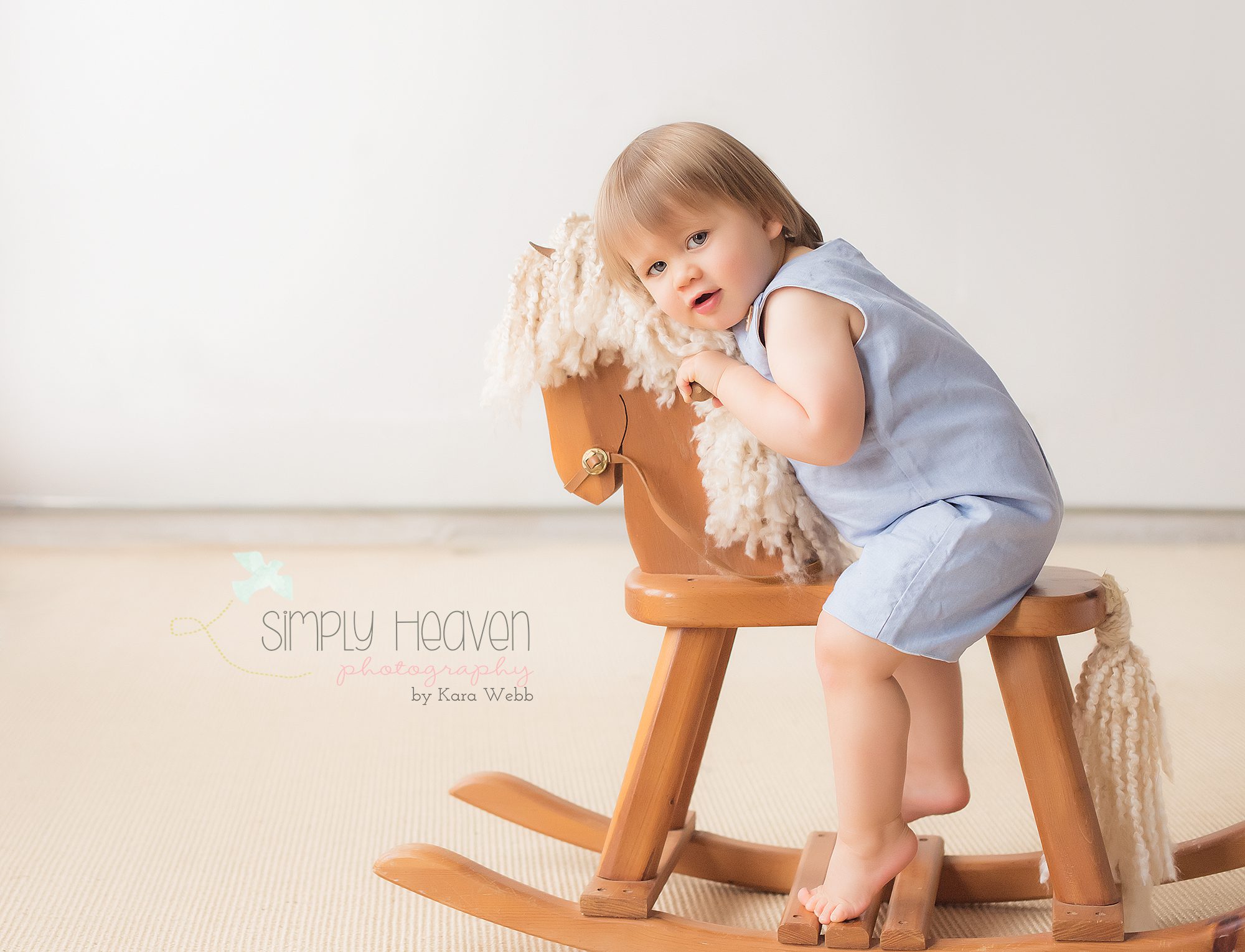 eighteen month old baby boy in a blue romper sitting on a wooden rocking horse