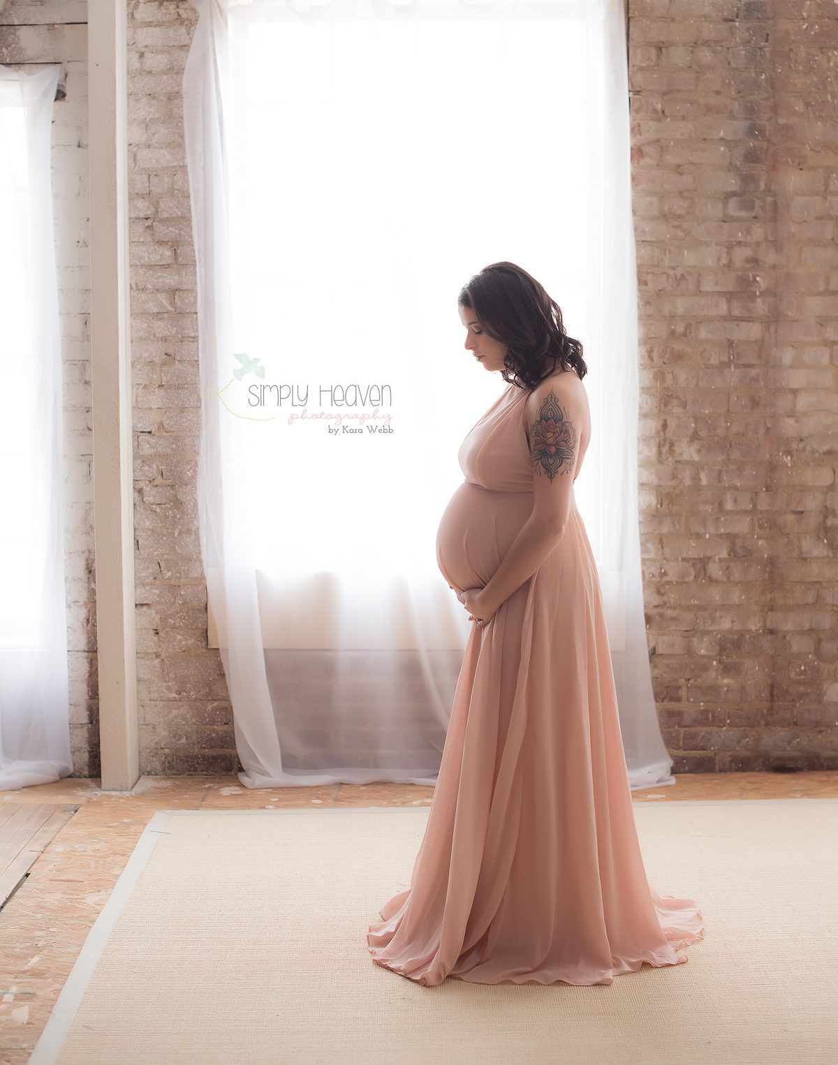 maternity pictures of a lady in a pink dress in front of brick wall and window