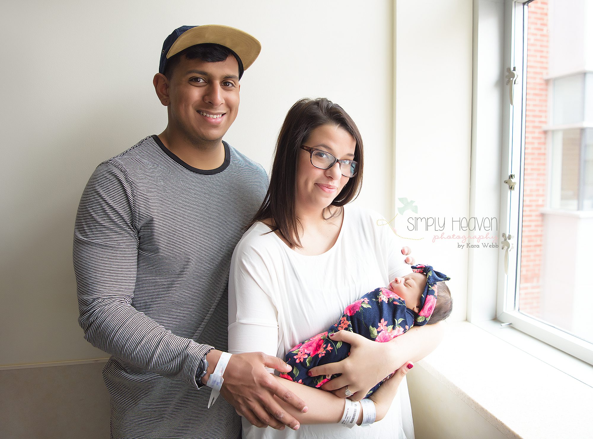 dad and mom holding her newborn baby girl in front of a hospital window
