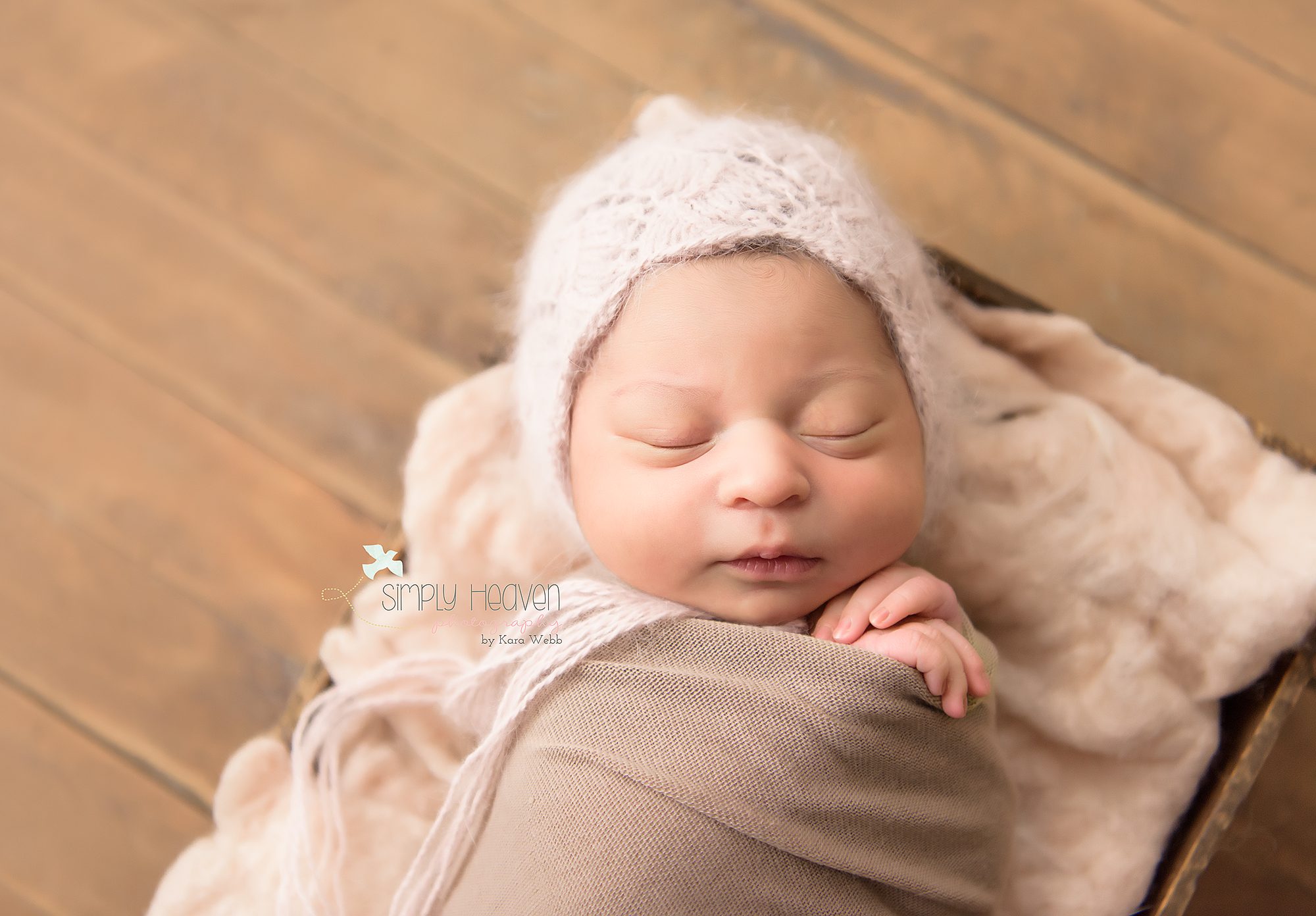 newborn baby girl swaddled in a fawn color with a soft pink angora bonnet with hands crossed