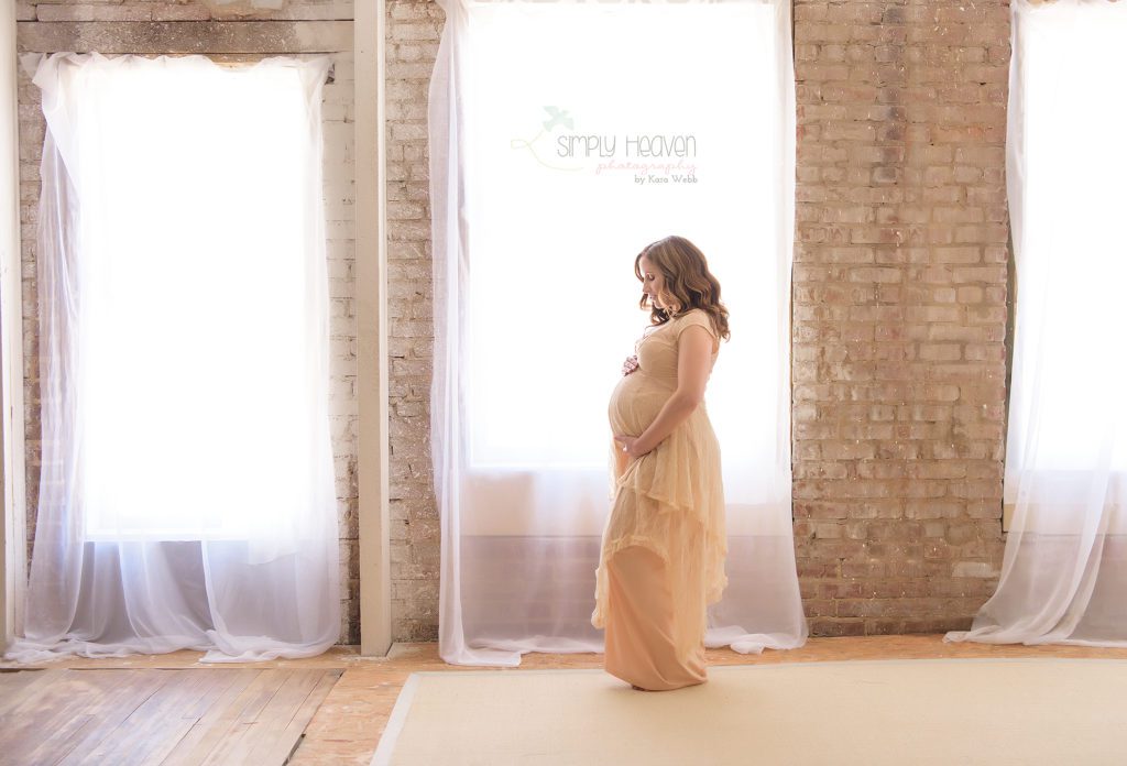 pregnant woman standing in front of a window during a maternity portrait session wearing a dress
